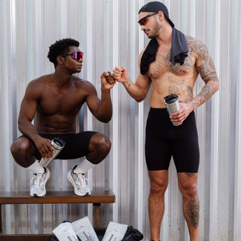 Zwei durchtrainierte Männer in Sportkleidung, einer sitzend, der andere stehend, stoßen sich vor einer Metallwand die Fäuste an, während sie Shakerflaschen halten. Beide tragen Sonnenbrillen, einer hat ein Handtuch über der Schulter und sichtbare Tattoos auf dem Arm.