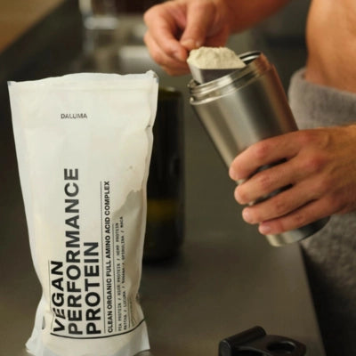 Eine Person schöpft veganes Hochleistungsproteinpulver aus einem Beutel mit der Aufschrift Vegan Performance Protein in eine Shakerflasche aus Edelstahl auf einer Küchenzeile.