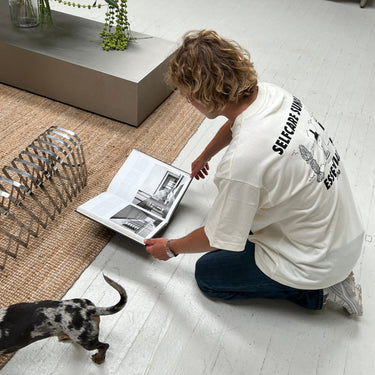 Eine Person mit gewelltem blondem Haar kniet auf einem weißen Boden und trägt das JOSEPH ATELIER X DALUMA SELFCARE SUNDAY TEE von DALUMA und Jeans. Ein kleiner schwarz-weißer Hund läuft in der Nähe in einem modernen, mit Pflanzen gefüllten Raum mit einem gewebten Teppich.