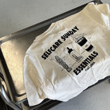 Das JOSEPH ATELIER X DALUMA SELFCARE SUNDAY TEE von DALUMA, ein T-Shirt aus nachhaltiger Bio-Baumwolle mit Oversize-Passform, trägt die Aufschrift "SELF-CARE SUNDAY ESSENTIALS DALUMA" und zeigt in schwarzer Schrift eine Kerze, eine Pflanze, ein Buch und ein Getränk.