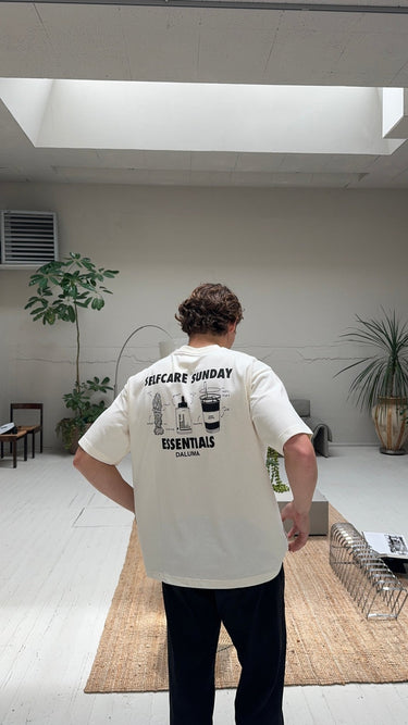 Eine Person mit lockigem Haar steht in einem Innenraum auf einem hellen Holzboden und trägt das DALUMA JOSEPH ATELIER X DALUMA SELFCARE SUNDAY TEE mit Illustrationen. Der minimalistische Raum ist mit Pflanzen und natürlichem Licht ausgestattet.