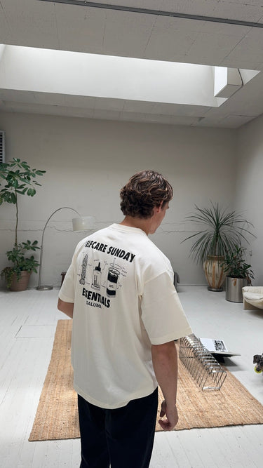 Eine Person mit braunem Haar steht in einem hellen, minimalistischen Raum und trägt das JOSEPH ATELIER X DALUMA SELFCARE SUNDAY TEE von DALUMA. Der Raum ist mit Topfpflanzen, einer Lampe und einem Juteteppich ausgestattet, die für eine nachhaltige, luftige Atmosphäre sorgen.