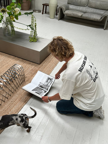 Eine Person, die das JOSEPH ATELIER X DALUMA SELFCARE SUNDAY TEE von DALUMA trägt, kniet auf einem weißen Boden und liest ein aufgeschlagenes Buch mit Schwarz-Weiß-Fotos. In dem hellen, modernen Raum mit minimalem Mobiliar und Pflanzen läuft ein kleiner Hund in der Nähe herum.
