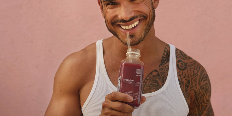Ein lächelnder Mann in einem weißen Tank-Top mit einer Tätowierung auf der Schulter hält eine Glasflasche mit rotem Saft und einem Strohhalm in der Hand und steht vor einer rosa Wand.