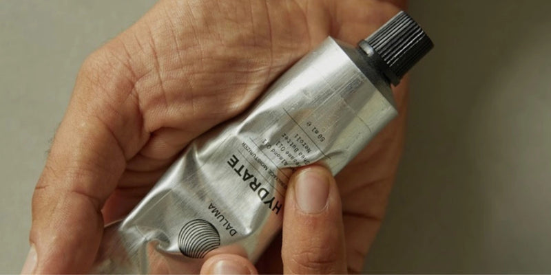 Eine Hand hält eine gequetschte silberne Tube mit der Aufschrift HYDRATE und einer schwarzen Kappe. Die Tube scheint eine Creme oder Lotion zu enthalten.