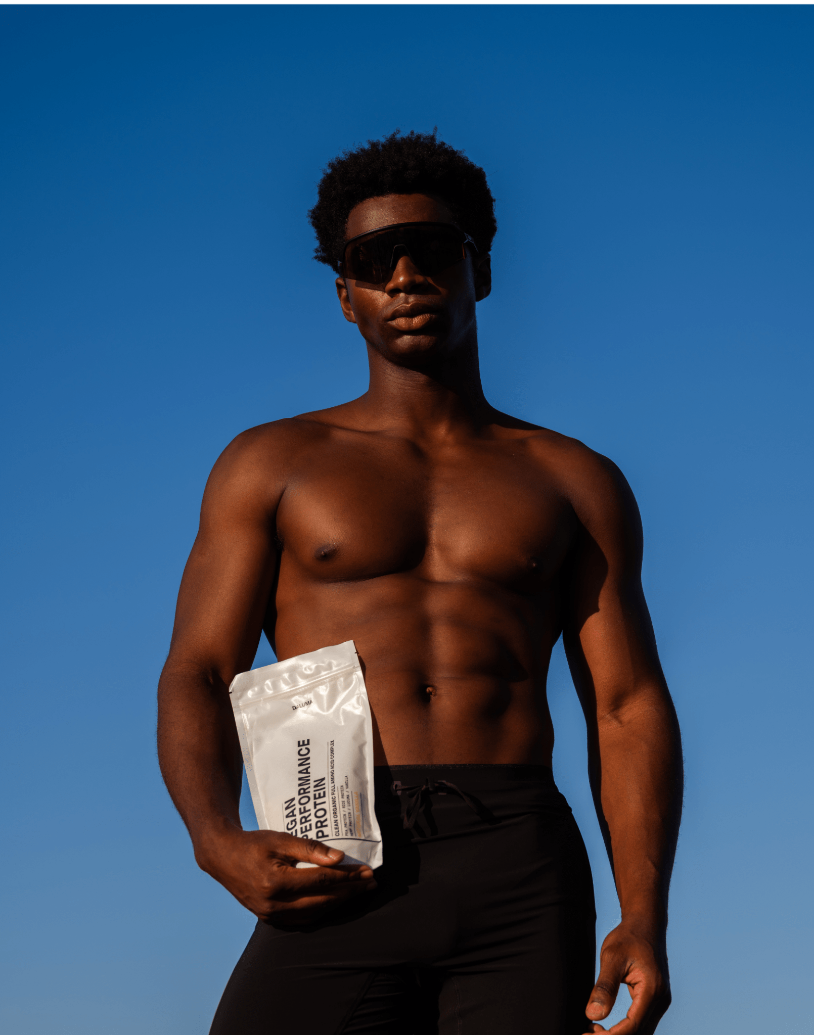 Ein fitter, selbstbewusster Mann mit schwarzer Sonnenbrille steht unter einem klaren, blauen Himmel und hält einen DALUMA PERFORMANCE DUO KAKAO Beutel in der Hand, der die Kraft von pflanzlichem Eiweiß für effektiven Muskelaufbau unterstreicht.