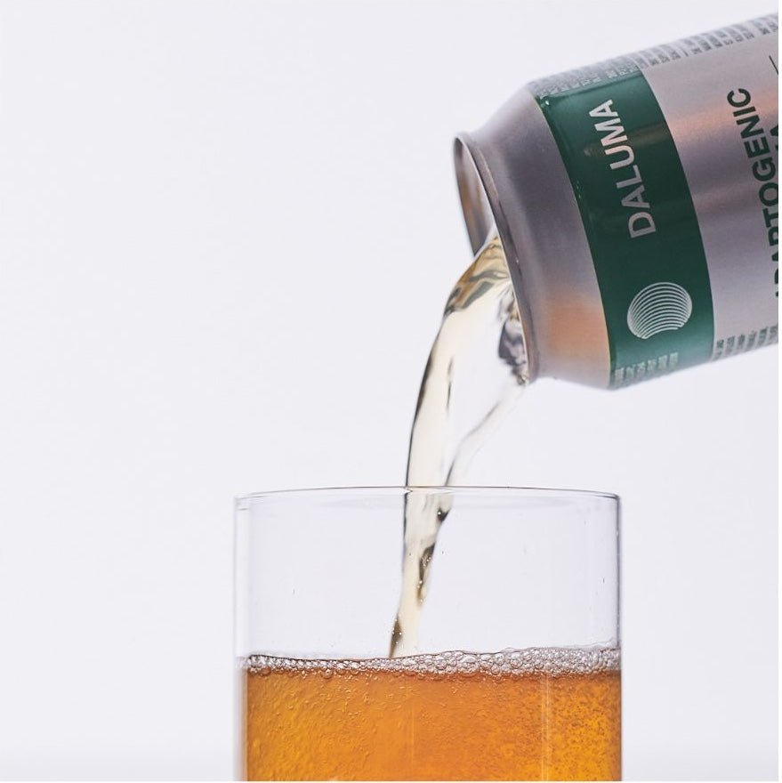 Eine Dose mit der Aufschrift DALUMA GLOW UP GREENS MARACUJA KOMBUCHA gießt ein hellbraunes, probiotikahaltiges Getränk in ein klares Glas vor weißem Hintergrund. Das Glas ist teilweise mit dem erfrischenden Kombucha gefüllt.