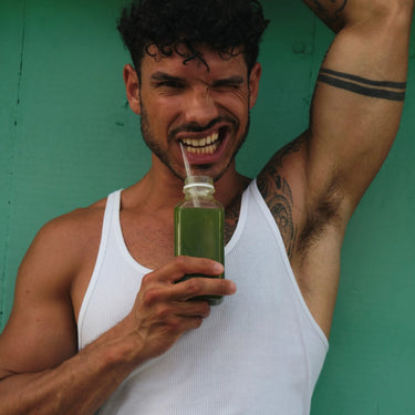 Ein Mann in einem weißen Tank-Top hält eine Flasche DALUMA GREEN JUICE PLUS SAFTKUR 5 TAGE mit einem Strohhalm, zwinkert und lächelt in die Kamera vor einem grünen Hintergrund - ideal für Ihre 5-tägige Saftkur.