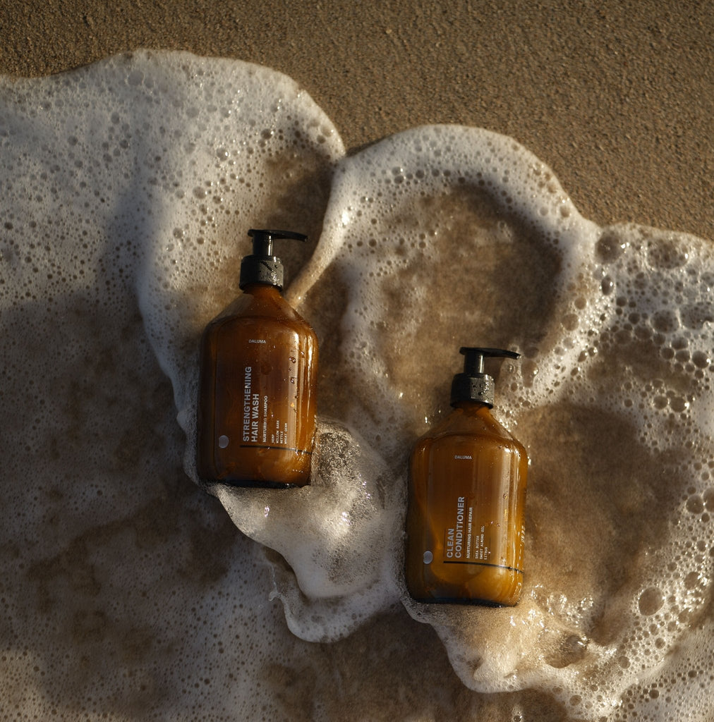 Zwei braune Pumpflaschen des Daluma-shop HAIR CARE DUO mit minimalistischen weißen Etiketten ruhen auf nassem Sand inmitten von Meeresschaum, perfekt positioniert am Ufer, um luxuriöses Haarwachstum zu unterstützen.