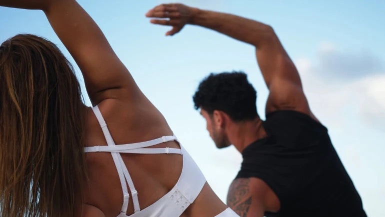 Zwei Personen, die im Freien in der Nähe des Ozeans trainieren und sich mit ausgestreckten Armen zur Seite strecken. Die Frau vorne trägt einen weißen Sport-BH und einen Rock, während der Mann hinter ihr ein schwarzes Hemd trägt. Der Himmel ist blau und teilweise bewölkt.