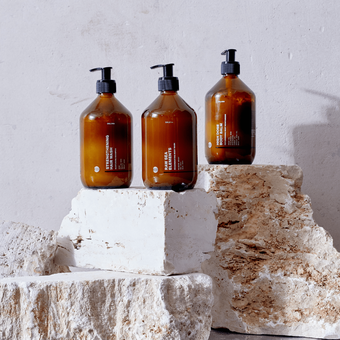 Drei bernsteinfarbene Pumpflaschen von DALUMA HIGH DOSE BODY BALM mit weißem Etikett stehen auf hellen Steinblöcken vor einer neutralen Wand - ideal für alle, die Falten reduzieren und ihre Hautbarriere stärken wollen.