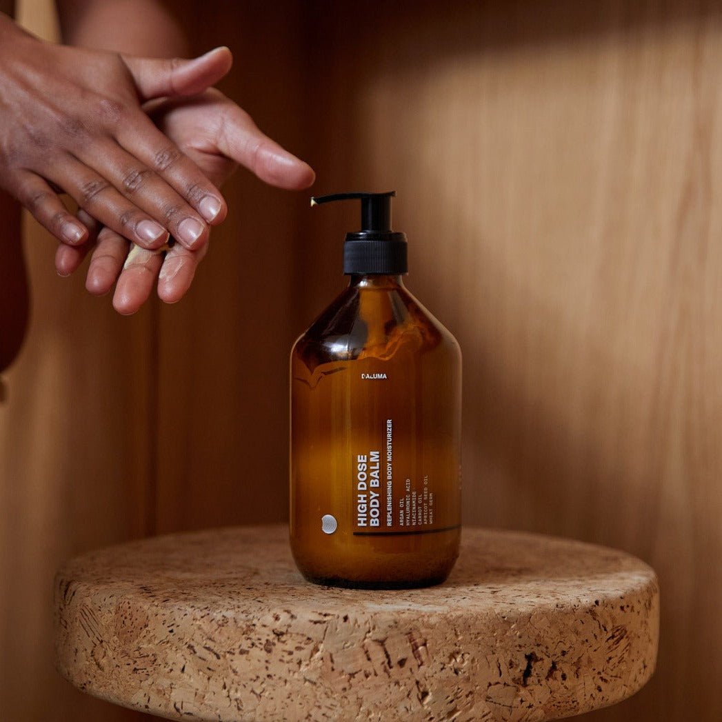 Eine Person reibt ihre Hände aneinander in der Nähe einer Pumpflasche aus braunem Glas mit der Aufschrift DALUMA SKIN NOURISHING DUO, die auf einer runden Korkfläche mit Holzuntergrund steht.