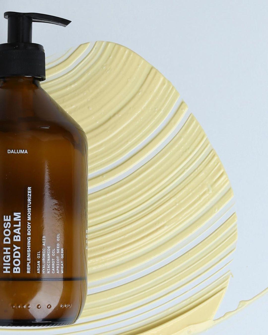 Eine braune Pumpflasche mit DALUMA HIGH DOSE BODY BALM liegt auf einer weißen Fläche mit einem Wirbel aus gelber Creme - ideal, um die Hautbarriere zu unterstützen und Falten zu reduzieren.