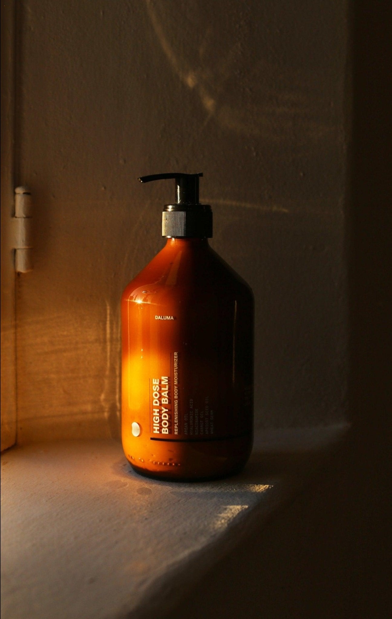 Die bernsteinfarbene Pumpflasche von DALUMA HIGH DOSE BODY BALM steht auf einer sonnenbeschienenen Fensterbank und wirft sanfte Schatten. Mit Arganöl formuliert, um die Hautbarriere zu stärken.
