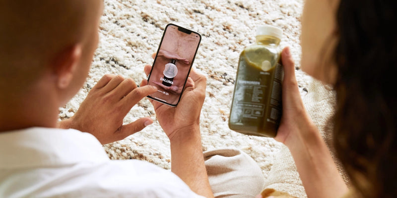 Zwei Personen sitzen eng beieinander auf einem Teppich. Eine hält ein Smartphone in der Hand, auf dem eine digitale Geldbörse mit einem Münzsymbol und dem Wort GUAMAN abgebildet ist. Die andere hält ein grünes Getränk in der Hand. Nur ihre Hände und teilweise ihre Gesichter sind zu sehen.