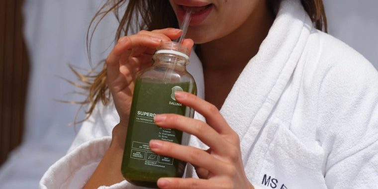 Eine Person in einem weißen Gewand trinkt grünen Saft aus einer Glasflasche mit Strohhalm. Der Fokus liegt auf ihren Händen, ihrem Mund und der Flasche, die ein Etikett mit Text trägt. Der Hintergrund ist unscharf.