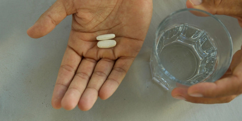 Eine Hand hält zwei weiße Tabletten, während die andere Hand ein Glas Wasser auf einer hellen Fläche hält.