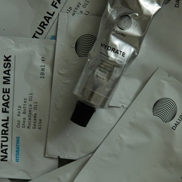 Mehrere DALUMA HYDRATING ESSENTIALS Produkte, darunter eine Metalltube und flache Päckchen mit der Aufschrift Natural Face Mask und Inhaltsstofflisten, sind auf einer wassergetränkten Fläche verstreut. Das DALUMA-Logo erscheint auf jedem Produkt.