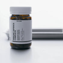 Eine braune Glasflasche mit der Aufschrift IMMUN DEFENCE COMPLETE KOMPLEX DOUBLE von DALUMA mit weißer Kappe und Etikett mit schwarzem Text steht auf einer weißen Fläche; im Hintergrund befindet sich ein verschwommenes Metalltablett.