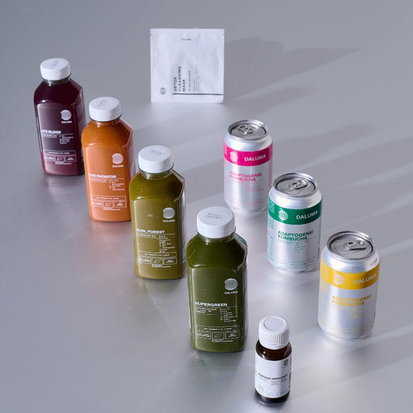 Eine Auswahl an Flaschensäften, Dosen und ein kleines Fläschchen, die auf einer grauen Fläche angeordnet sind, jeweils mit bunten Etiketten in Grün-, Orange-, Lila-, Rosa-, Gelb- und Weißtönen. Auf der Rückseite befindet sich ein weißes Päckchen.