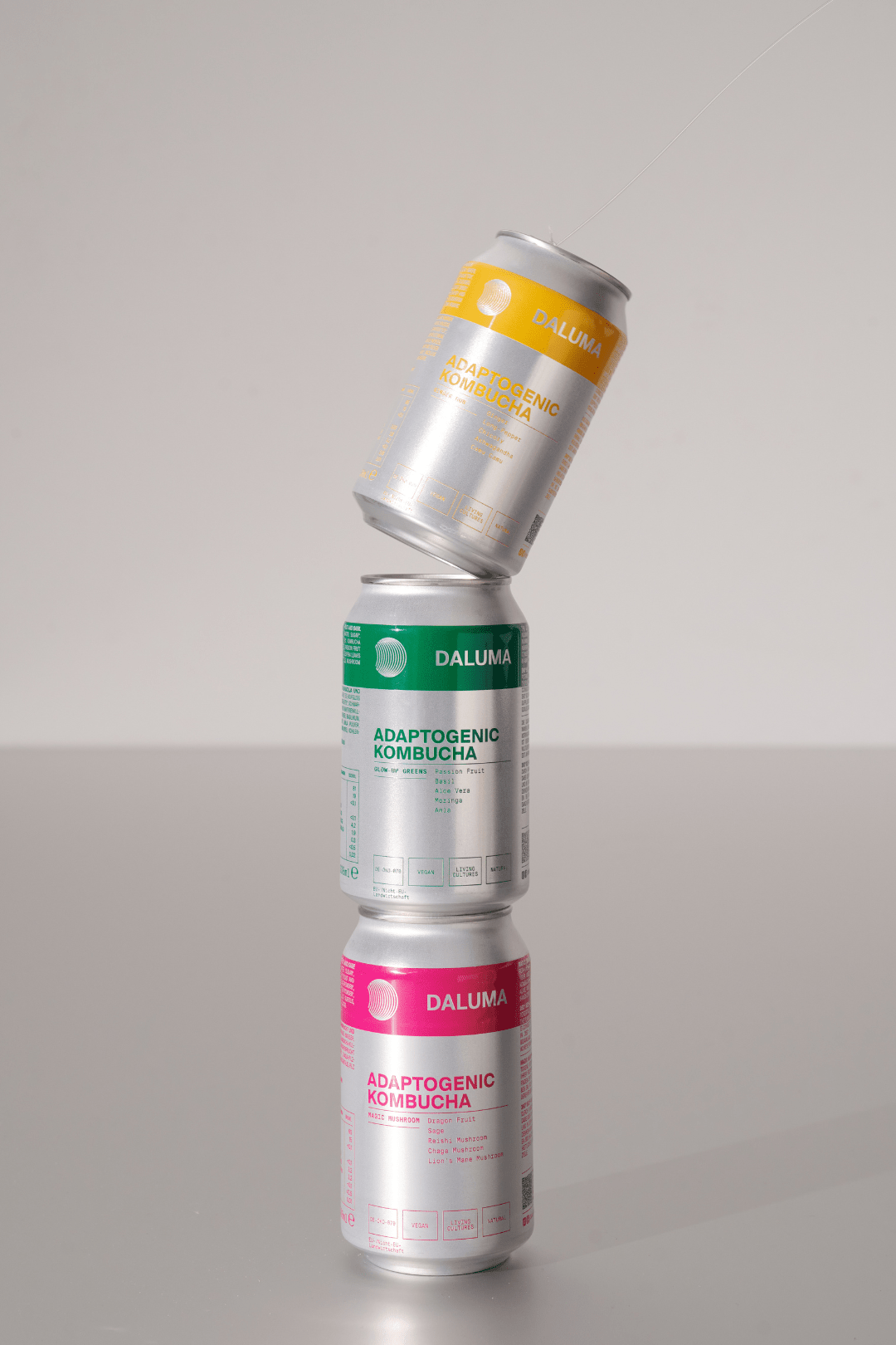 Drei Dosen DALUMA GLOW UP GREENS MARACUJA KOMBUCHA, voll mit probiotischen Kulturen für die Darmgesundheit, sind übereinander gestapelt - grün und rosa auf der Unterseite und eine gelbe Dose, die diagonal auf der Oberseite balanciert, vor einem schlichten Hintergrund.