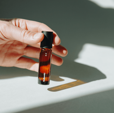 Eine Hand hält eine kleine Braunglasflasche mit einer schwarzen Kappe mit der Aufschrift "NULLPUNKT GRADE 0.2 RELEASE" von Nullpunkt. Die Flasche wirft im hellen Sonnenlicht einen Schatten auf eine weiße Fläche. Es ist keine Produktbeschreibung vorhanden.