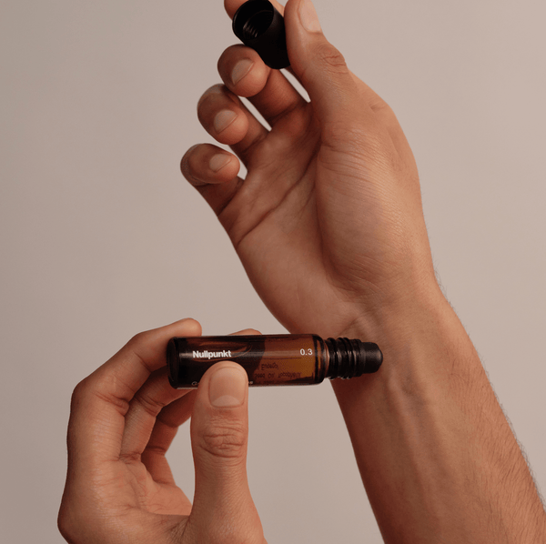 Eine Person hält einen kleinen Roll-on-Flakon aus Braunglas mit der Aufschrift NULLPUNKT GRADE 0.3 REFOCUS von Nullpunkt in der Hand und schraubt den schwarzen Verschluss mit einer Hand ab, während sie den Flakon vor einem minimalistischen beigen Hintergrund hält.