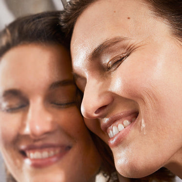 Eine Person mit kurzen braunen Haaren lächelt, ihre Wangen spiegeln vor einem Spiegel strahlende Haut und Freude wider - und zeigen die Vorteile von DALUMA PREBIOTIC PURIFYING GESICHTSSERUM für einen gesunden, strahlenden Teint.