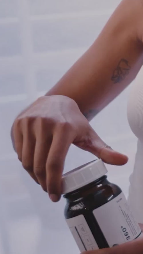 Eine Person in einem weißen ärmellosen Oberteil, die eine geometrische Oberarmtätowierung zeigt, öffnet eine Daluma 360 DAILY ESSENTIALS SUPPLEMENT DOUBLE Flasche mit weißem Deckel.
