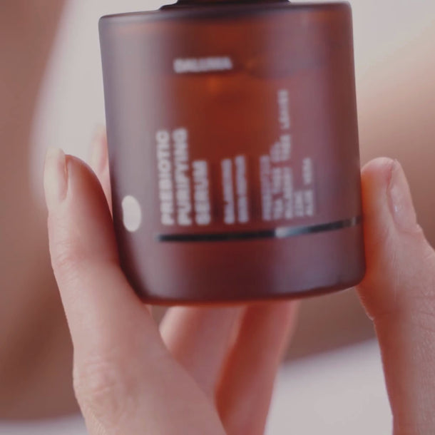 Eine Hand hält einen braunen, gefrosteten Tiegel mit der Aufschrift Prebiotic Purifying Cream, wobei der Text teilweise sichtbar ist; der Hintergrund ist leicht unscharf.