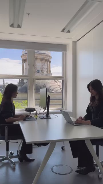 Zwei Frauen sitzen sich an einem weißen Schreibtisch in einem modernen Büro gegenüber und arbeiten an Laptops. Große Fenster hinter ihnen geben den Blick frei auf ein historisches Gebäude mit Kuppeldach unter blauem Himmel.