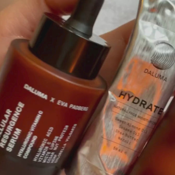 Eine Hand hält eine braune Glastropfflasche mit der Aufschrift Cellular Resurgence Serum neben einer Metalltube mit der Aufschrift Hydrate der Marke Daluma.