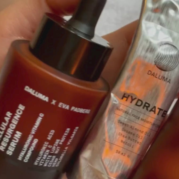 Eine Hand hält eine braune Glastropfflasche mit der Aufschrift Cellular Resurgence Serum neben einer Metalltube mit der Aufschrift Hydrate der Marke Daluma.