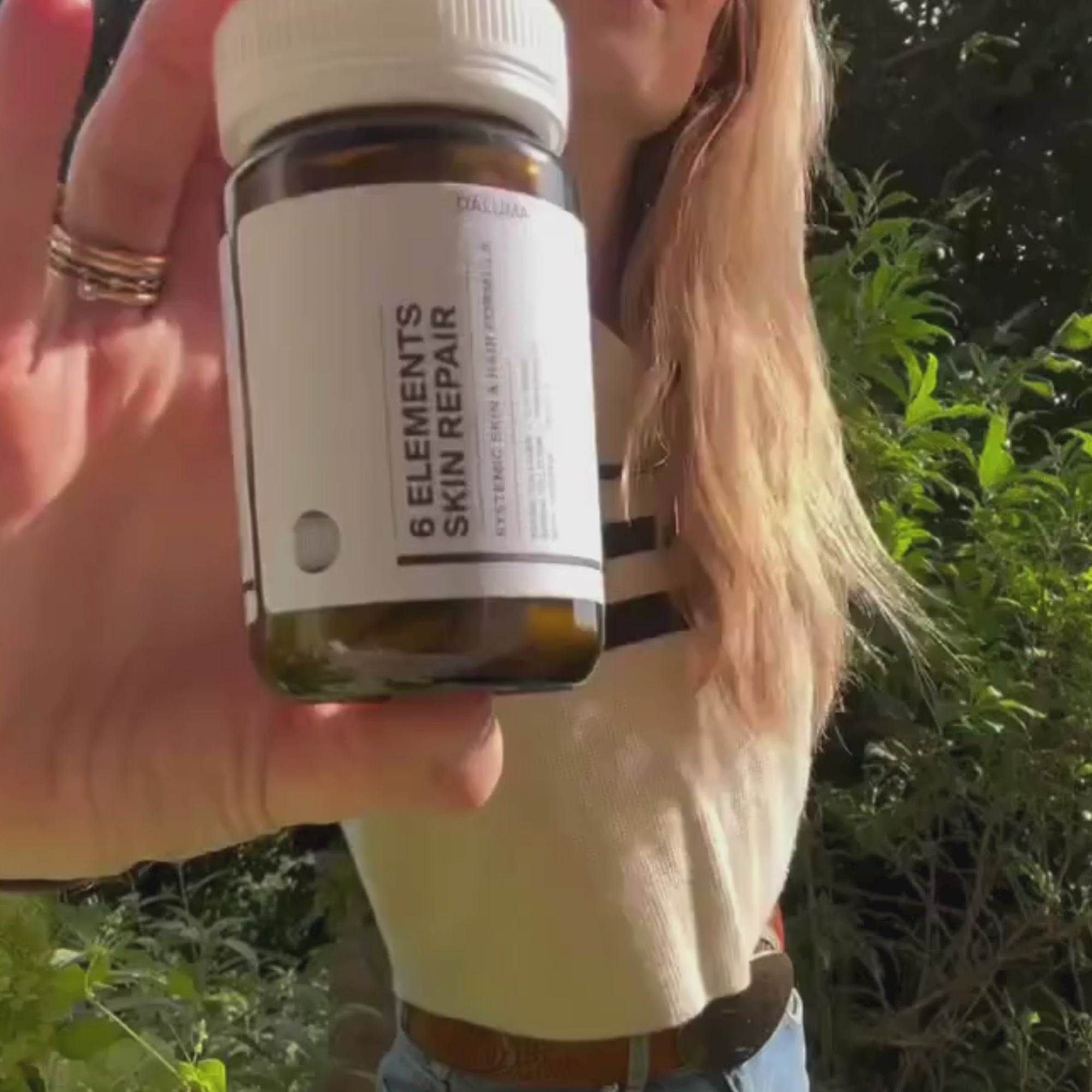 Eine Frau mit langen blonden Haaren hält eine braune Flasche mit der Aufschrift 6 Elements Skin Repair in die Höhe, im Hintergrund grünes Laub. Der Fokus liegt auf der Flasche in ihrer Hand.
