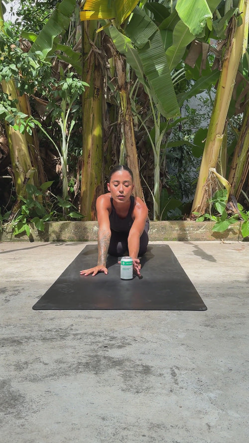 Eine Frau praktiziert Yoga im Freien auf einer schwarzen Matte und greift nach einer Dose Daluma DAILY KOMBUCHA BOOSTER PACKAGE, umgeben von großen grünen Bananenpflanzen. Sie trägt ein schwarzes Tanktop und Leggings.
