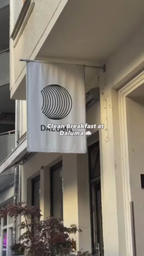 Ein weißes Schild mit einem kreisförmigen schwarzen Linienmuster hängt vor einem Gebäude. Der Schriftzug Clean Breakfast at Daluma ist über das Bild gelegt. In der unteren linken Ecke ist etwas Laub zu sehen.