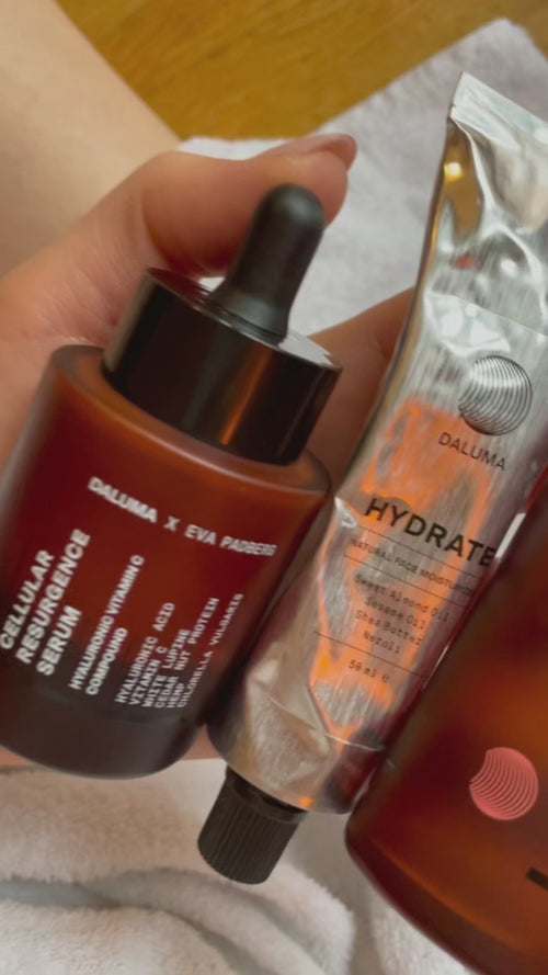 Eine Hand hält eine braune Glastropfflasche mit der Aufschrift "Cellular Resurgence Serum" und eine Metalltube mit der Aufschrift "Hydrate". Beide Hautpflegeprodukte sind von der Marke DALLUMA. Im Hintergrund sind ein Handtuch und eine Holzfläche zu sehen.