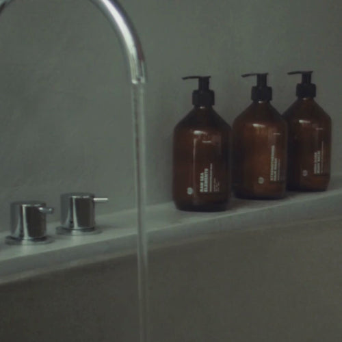 Eine Nahaufnahme eines Badewannenhahns mit fließendem Wasser neben drei braunen Daluma HAIR CARE DUO Pumpflaschen auf dem Wannenrand vor einer grauen Wand.