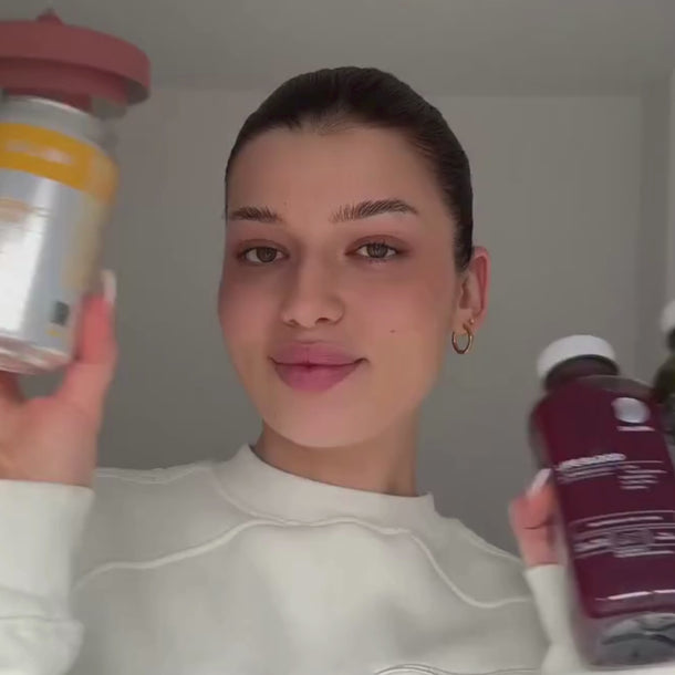 Eine Frau mit dunklem Haar und weißem Oberteil hält zwei Flaschen mit Daluma-Produkten in die Höhe und lächelt sanft in die Kamera vor einem schlichten, hellen Hintergrund.