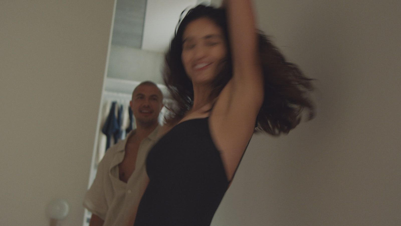Eine Frau in einem schwarzen Kleid lächelt und wirbelt in einem Flur herum, während eine andere Person im Hintergrund steht, sie anschaut und lächelt. Die Szene wirkt fröhlich und unbeschwert.