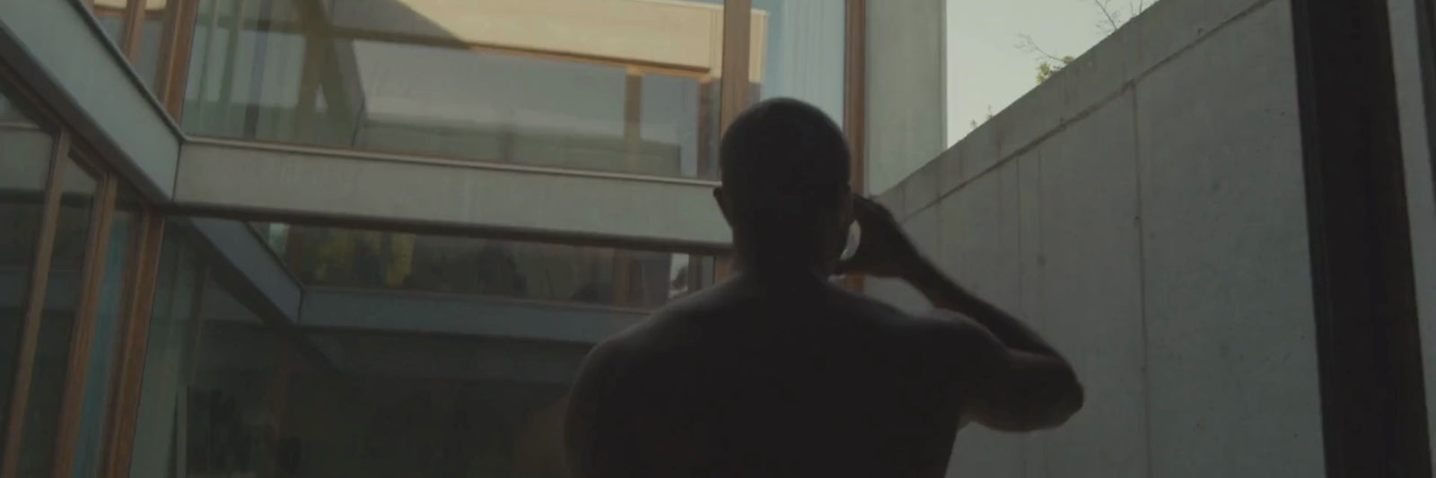 Eine Person mit rasiertem Kopf steht ohne Hemd in einem Innenraum vor großen Glasfenstern und Betonwänden und hält sich eine Tasse an den Mund in einem modernen, minimalistischen Gebäude mit natürlichem Licht.