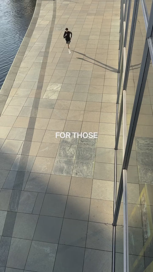 Eine Person geht allein auf einem breiten, gefliesten Gehweg neben Wasser und einem Glasgebäude, wobei die Worte FÜR DIE in der Mitte des Bildes stehen.