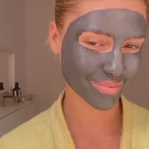 Eine Frau mit einer grauen Gesichtsmaske und einem gelben Bademantel lächelt sanft. In einem Regal im Hintergrund stehen Hautpflegeprodukte.