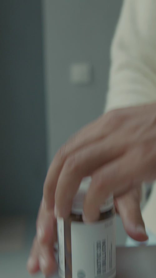 Eine Nahaufnahme einer Hand, die nach einer weißen Flasche mit einem Etikett greift, möglicherweise ein Medikament oder ein Nahrungsergänzungsmittel, vor einem unscharfen Innenhintergrund.