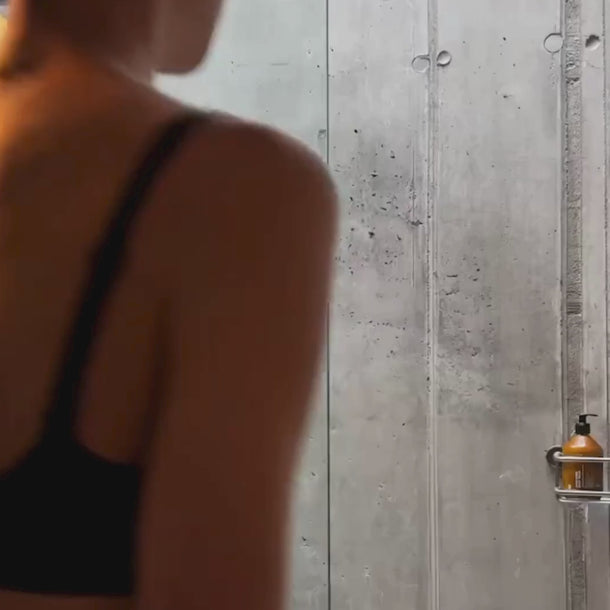 Eine Person in einem schwarzen BH steht vor einer Betonmauer, rechts davon ist eine braune Daluma BODY CARE DUO Pumpflasche auf einem metallenen Duschwagen zu sehen.