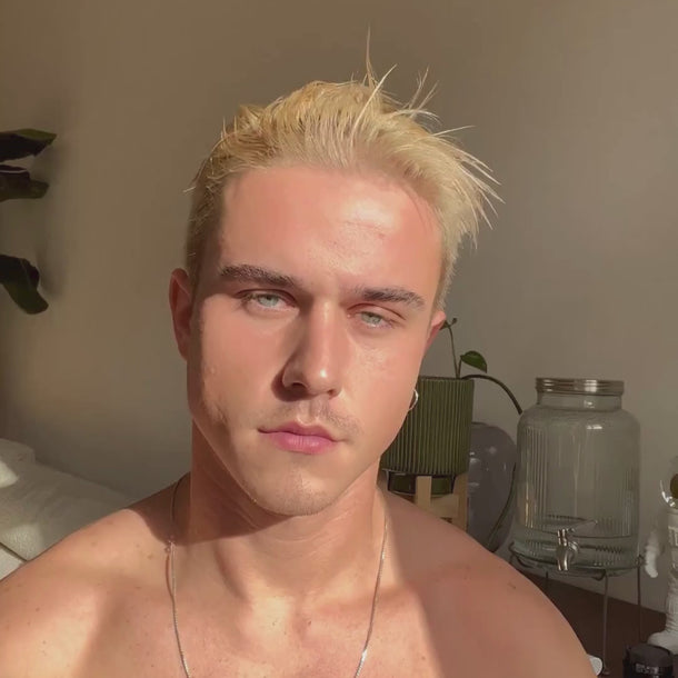 Eine hemdsärmelige Person mit kurzen blonden Haaren gibt Daluma BODY CARE DUO Lotion aus einer Pumpflasche in die Hand. Sie sitzt in einem Innenraum, die Sonne scheint auf ihr Gesicht und im Hintergrund sind Pflanzen zu sehen.