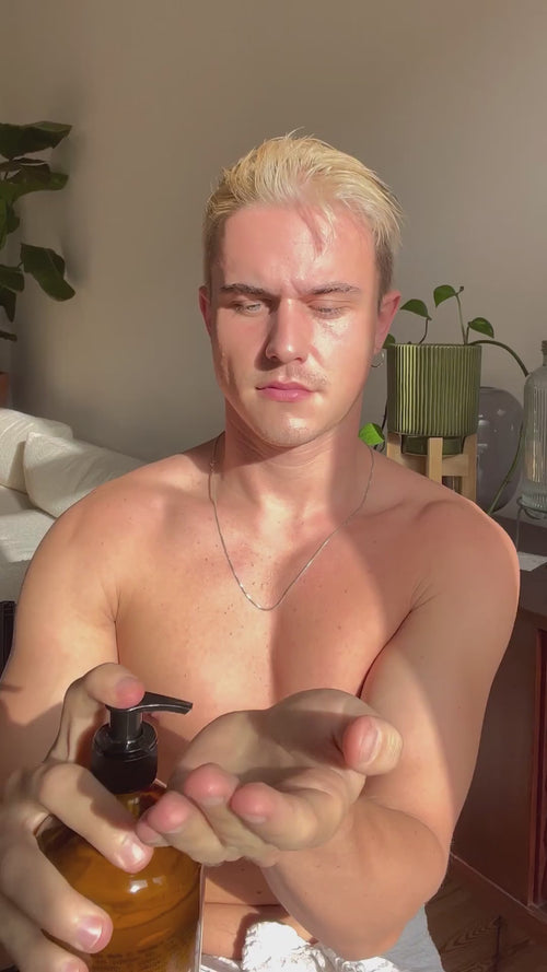 Eine hemdsärmelige Person mit kurzen blonden Haaren gibt Daluma BODY CARE DUO Lotion aus einer Pumpflasche in die Hand. Sie sitzt in einem Innenraum, die Sonne scheint auf ihr Gesicht und im Hintergrund sind Pflanzen zu sehen.