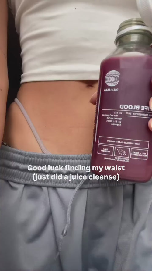 Eine Person in grauer Jogginghose und weißem Oberteil hält eine Flasche mit dunklem Saft. Der Text auf dem Bild lautet: "Viel Glück bei der Suche nach meiner Taille (habe gerade eine Saftkur gemacht)". Ihre Taille und der entblößte Unterwäscheträger sind sichtbar.