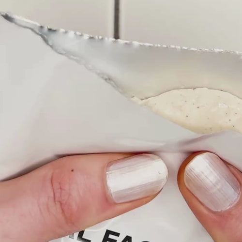 Eine Hand mit weiß lackierten Nägeln hält eine teilweise geöffnete weiße Packung Daluma DEEP HYDRATION FEUCHTIGKEITSMASKE, in der eine cremefarbene beigefarbene Maske zum Vorschein kommt.