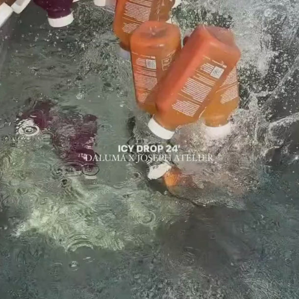 Mehrere orangefarbene Flaschen mit weißen Verschlüssen, die mit Daluma CLEANSE DAY beschriftet sind, plätschern ins Wasser - einige untergetaucht, andere mitten im Fall - und sind von Blasen und Wellen umgeben. Der Text auf dem Bild lautet "ICY DROP 24' DALUMA X JOSEPH ATELIER".